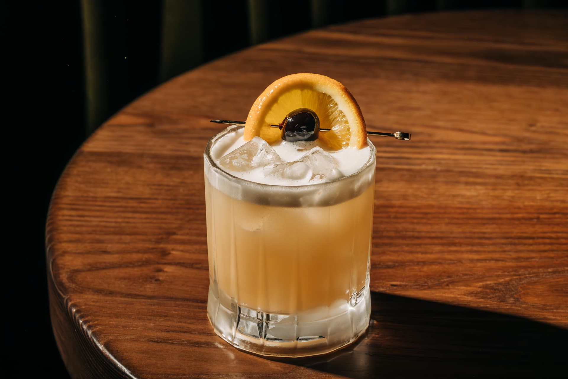A frothy cocktail served in a short glass on a wooden table, garnished with an orange slice and a cherry.