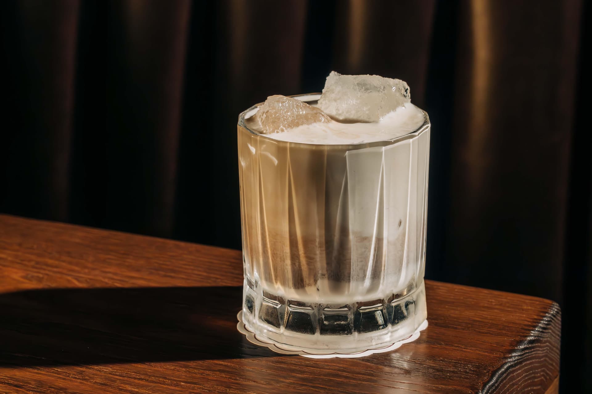 A glass filled with a creamy white Russian cocktail on a wooden table, topped with ice cubes.