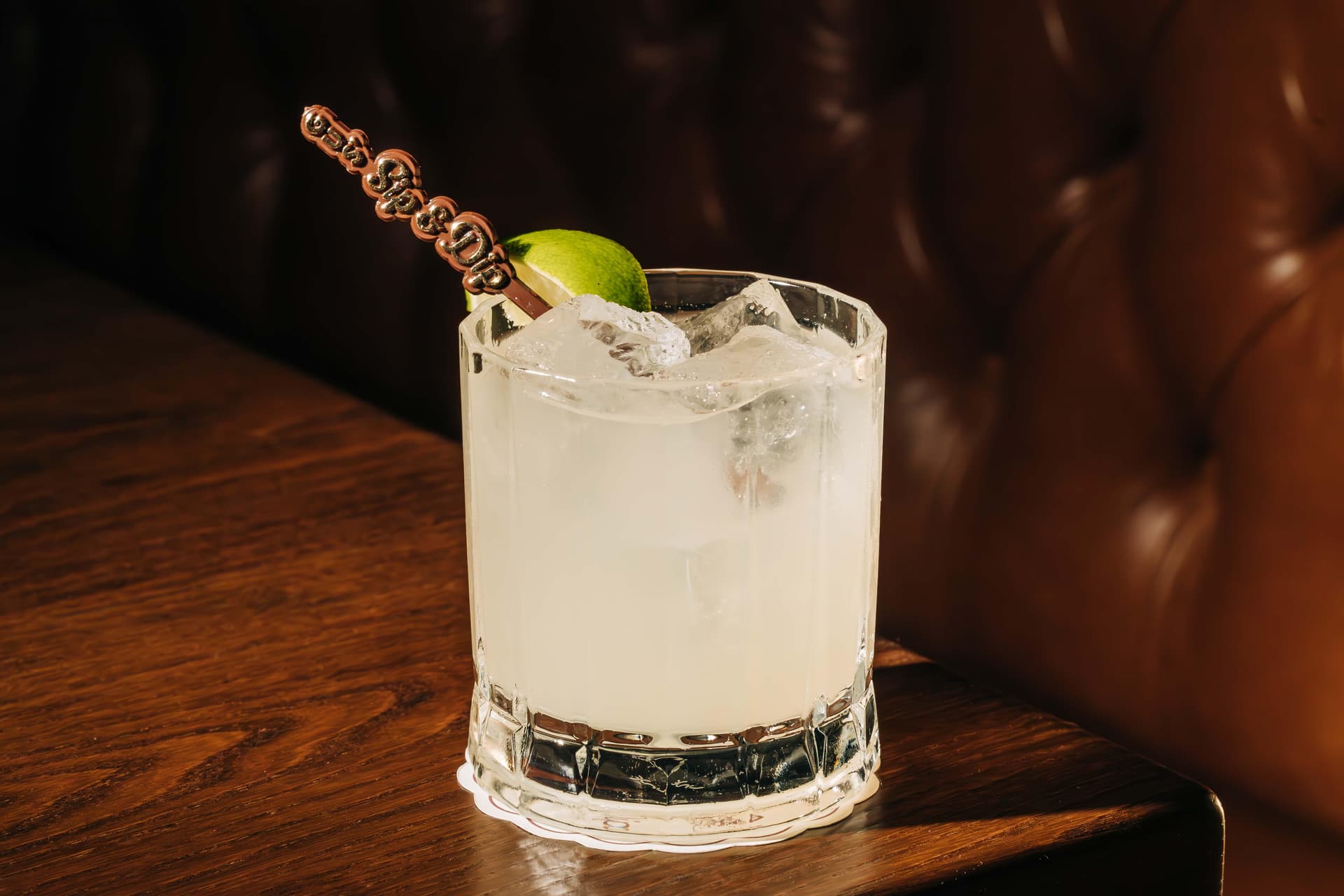 A frosted glass filled with a clear cocktail on ice, garnished with a lime wedge and a stirrer, placed on a wooden table. The background shows dark, tufted upholstery.