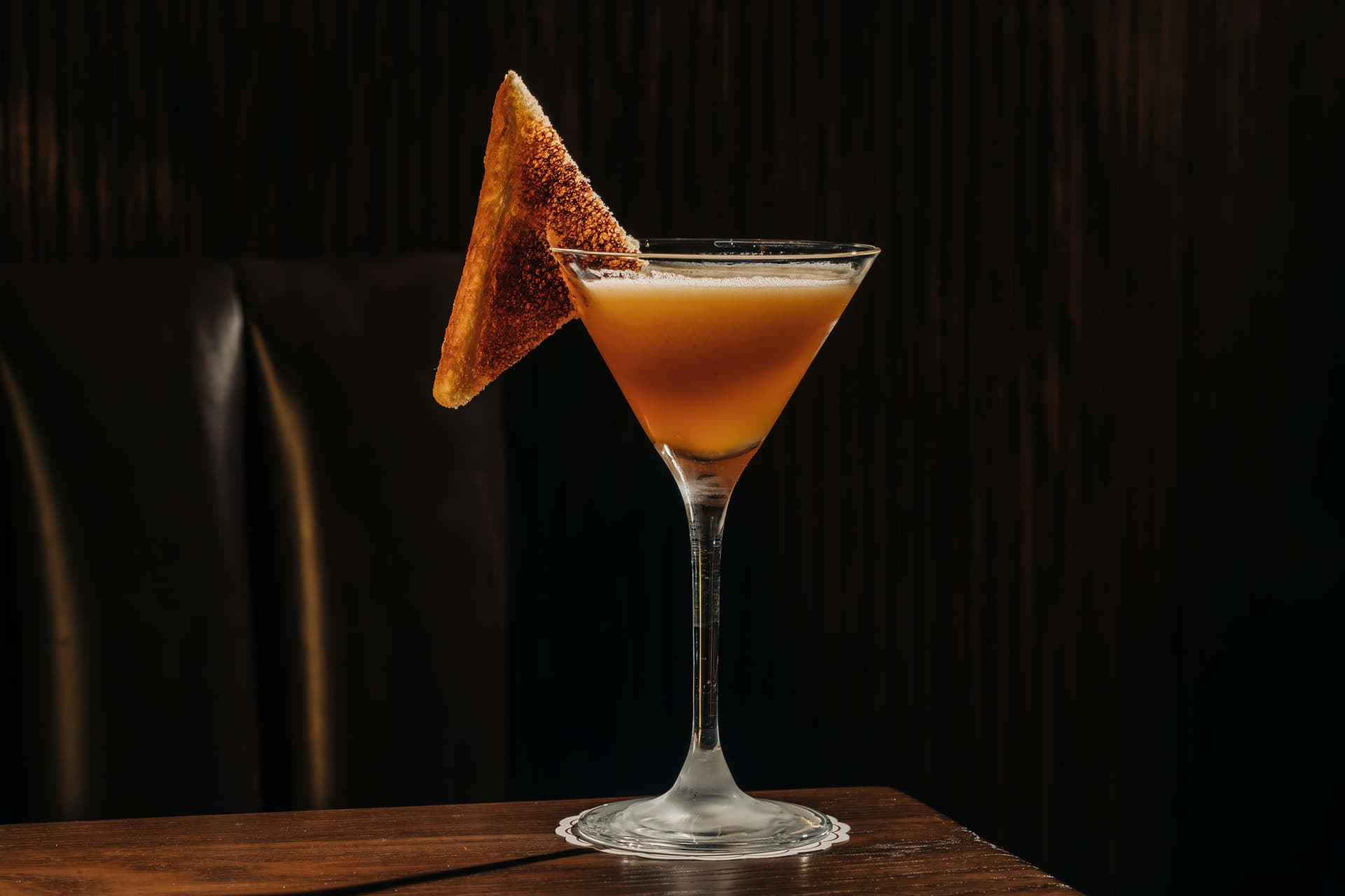 A martini glass filled with an orange drink, garnished with a triangular slice of toast, sits on a wooden table against a dark background.