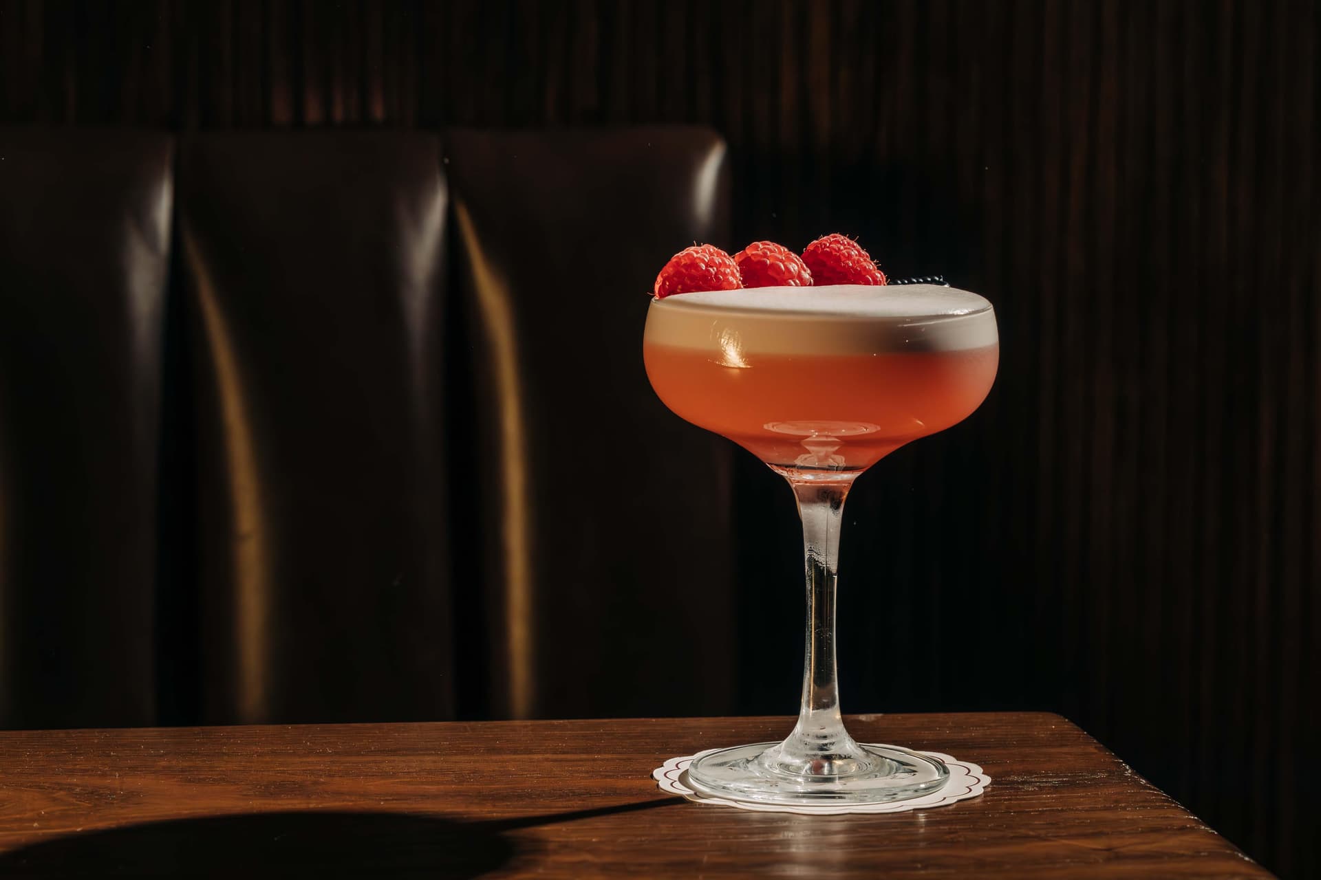A cocktail glass filled with a pink drink topped with foam and three raspberries, placed on a wooden table with a dark cushioned background.