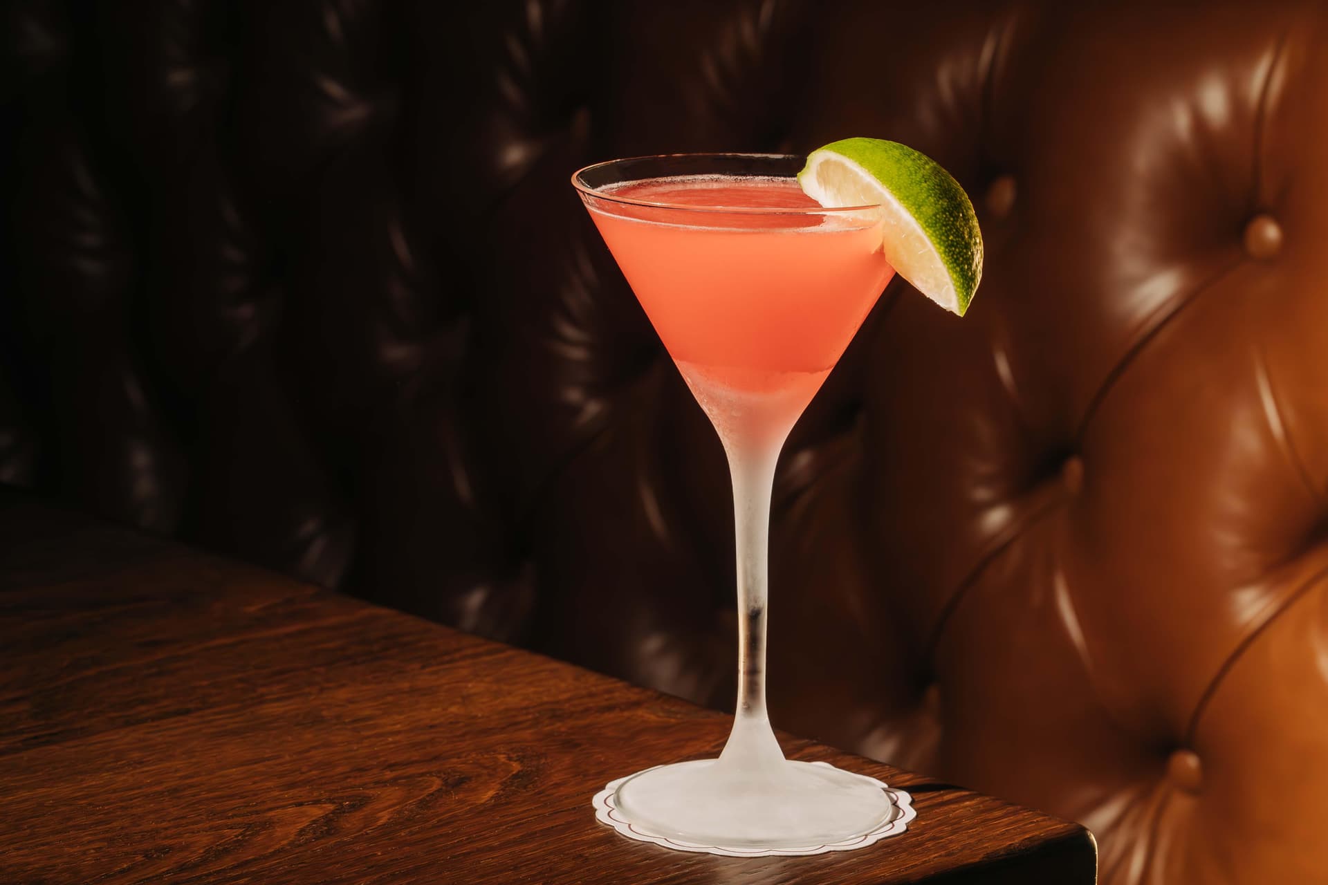 A pink cocktail in a martini glass with a lime wedge on the rim, placed on a wooden table with a tufted leather background.