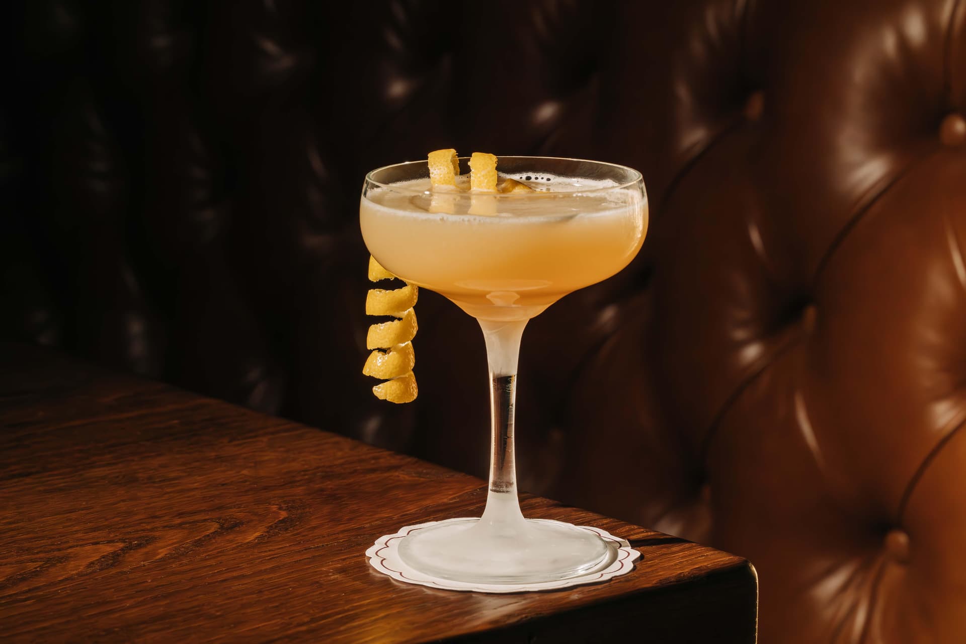 A coupe glass filled with a light amber cocktail, garnished with a lemon twist, sits on a wooden table. The background features dark brown, tufted leather upholstery.