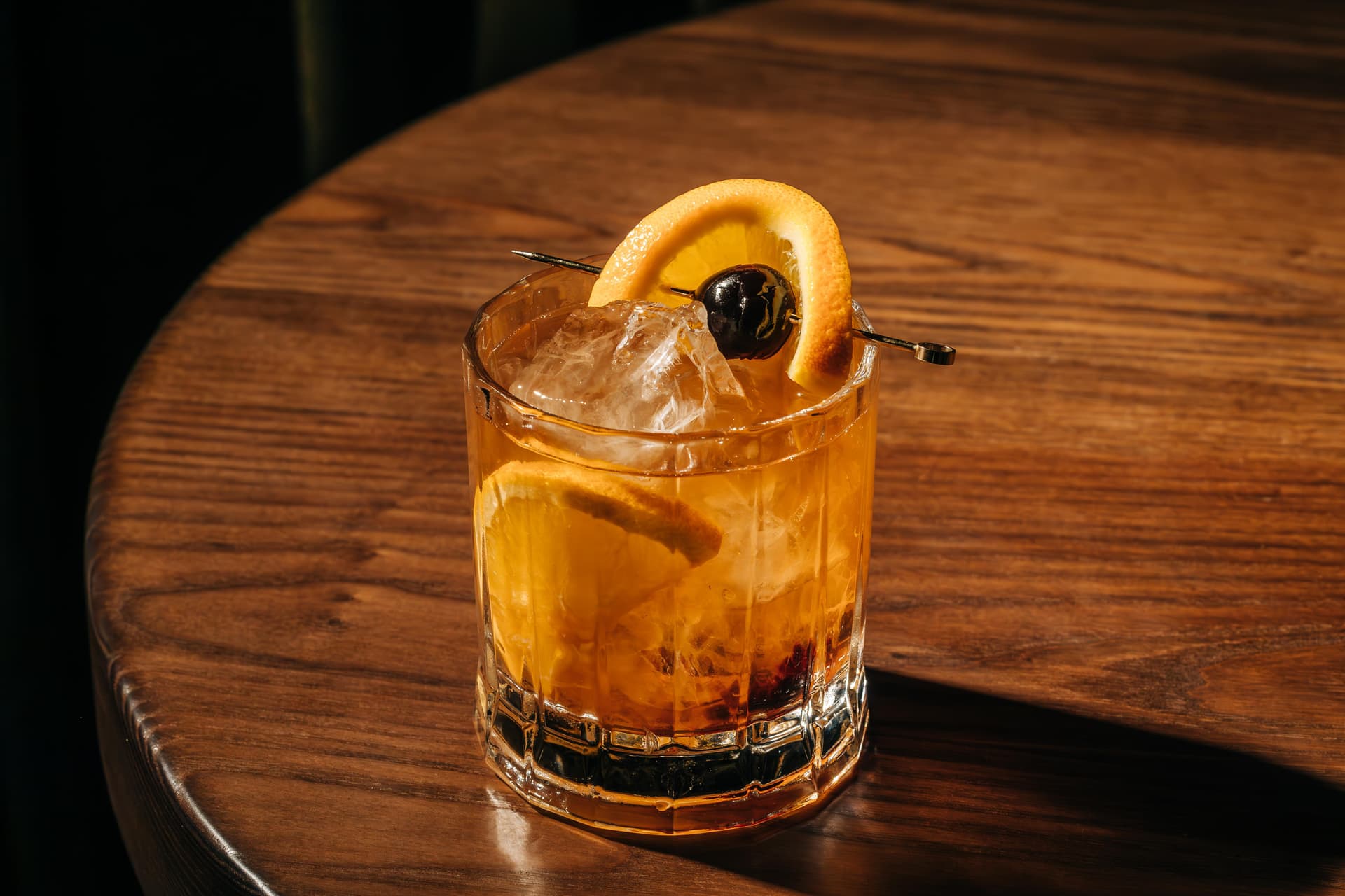 A glass of an Old Fashioned cocktail sits on a wooden table. The drink contains ice and is garnished with an orange slice and a cherry.