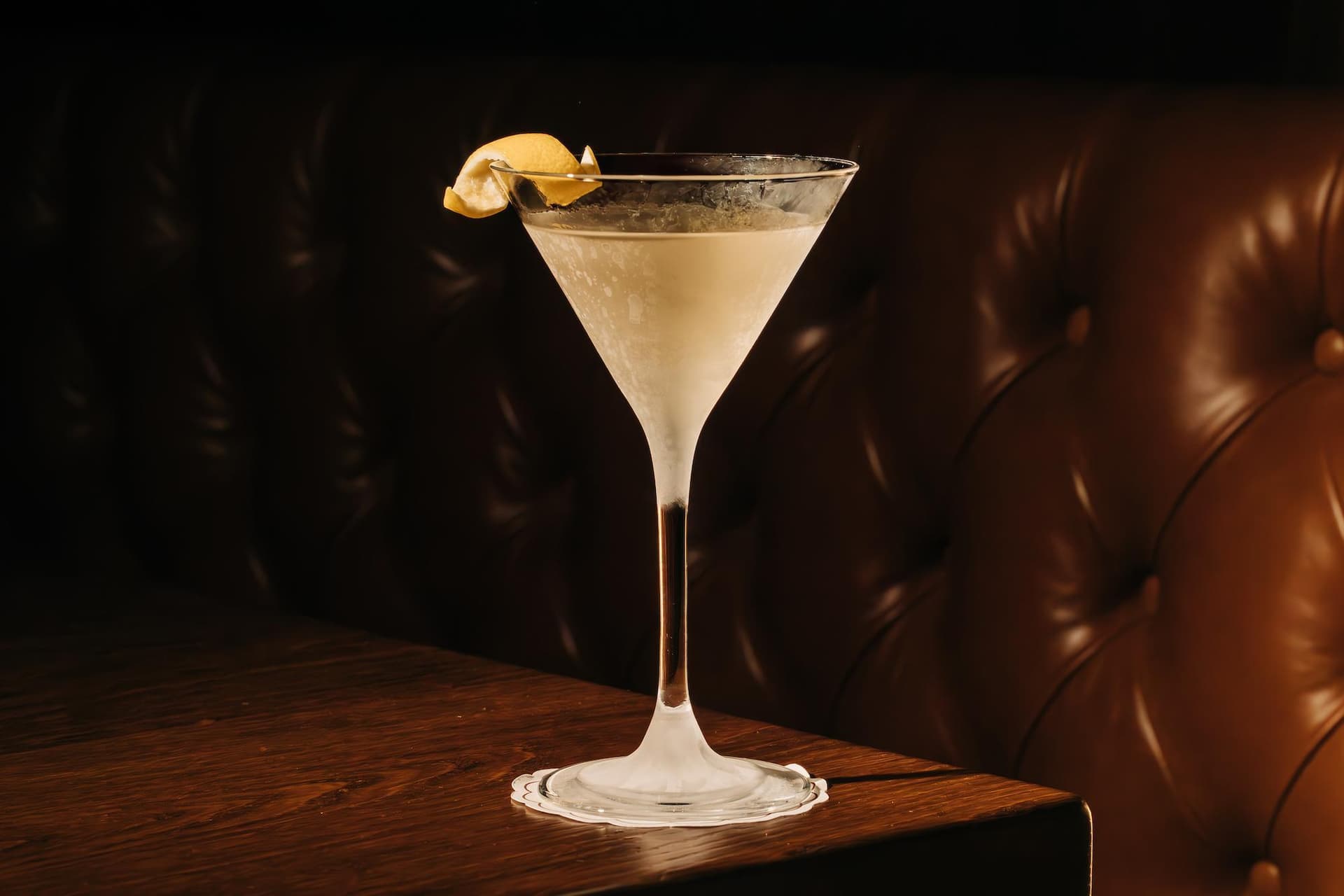 A martini glass with a light-colored cocktail garnished with a lemon twist, set on a wooden table against a tufted leather background.