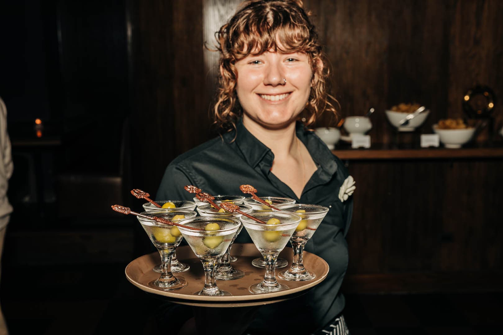 A server at a bar is smiling and holding a tray of martinis, each garnished with an olive.