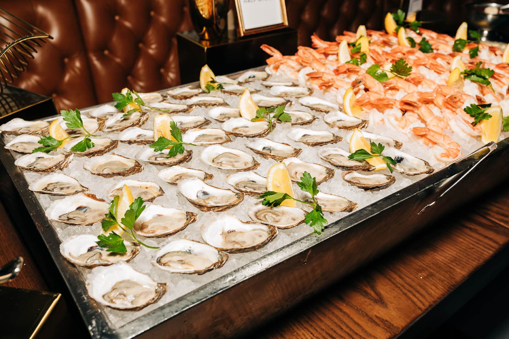 An array of fresh oysters and shrimp served on ice, garnished with lemon wedges and parsley, displayed on a table with a leather couch in the background.