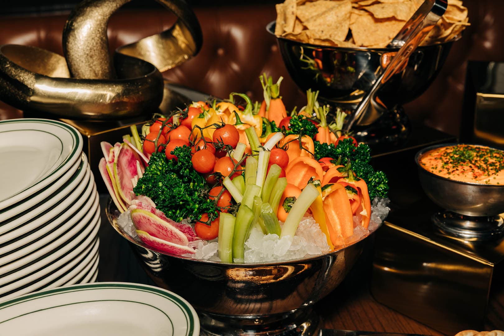 A variety of fresh vegetables including carrots, tomatoes, and celery, arranged in a serving bowl on ice, alongside plates and a bowl of chips, set up for a buffet.