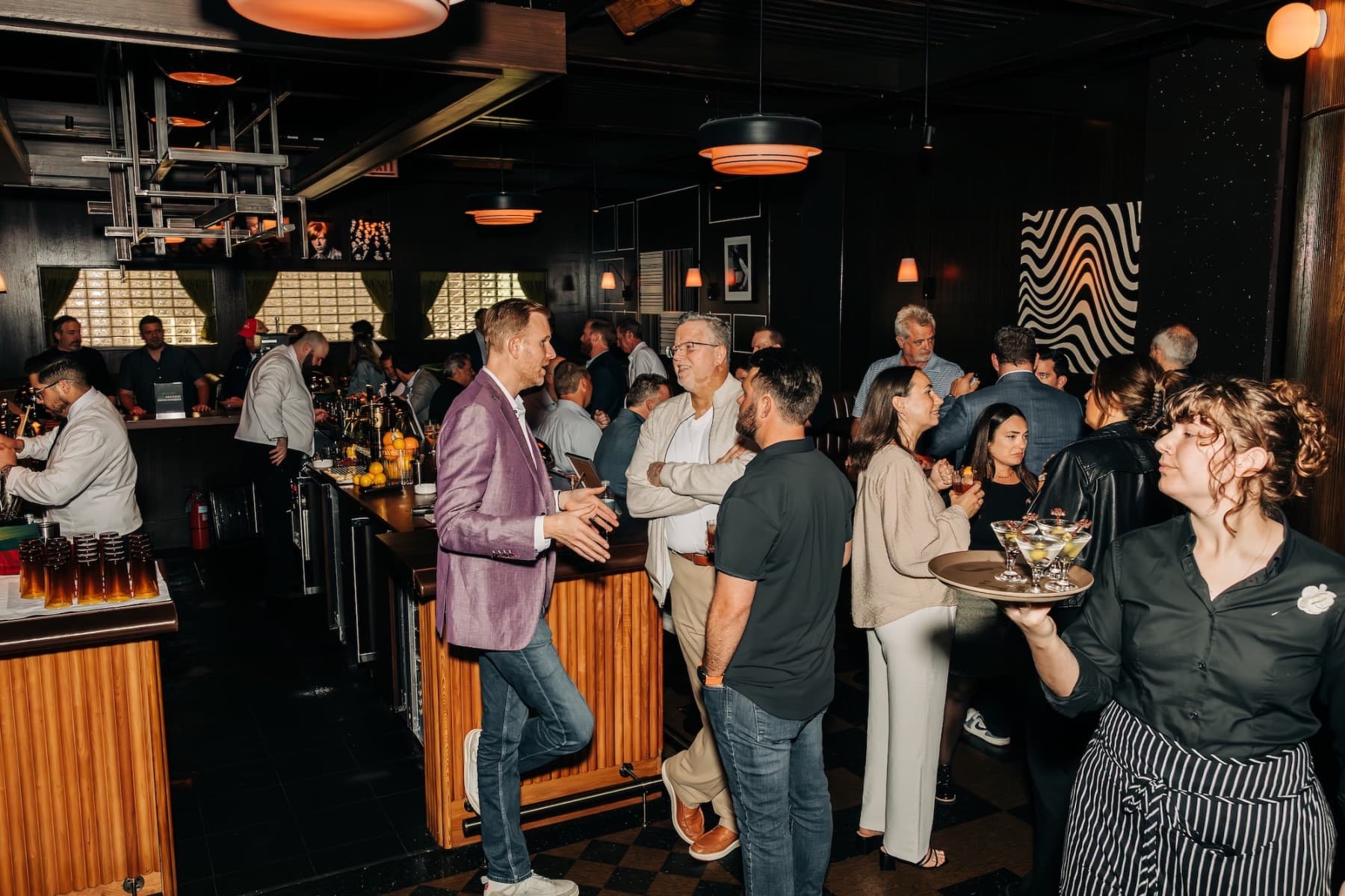 Busy social gathering at a bar with individuals engaged in conversations and a server carrying drinks.