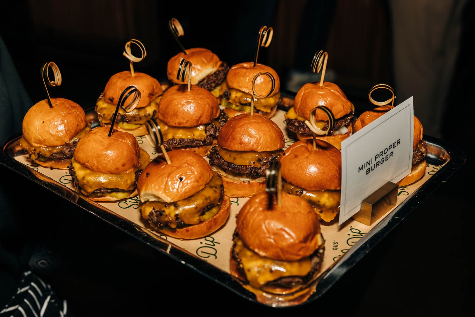 Tray of mini cheeseburgers served at an event, each garnished with a pickle and held together by a small skewer, labeled with a sign.
